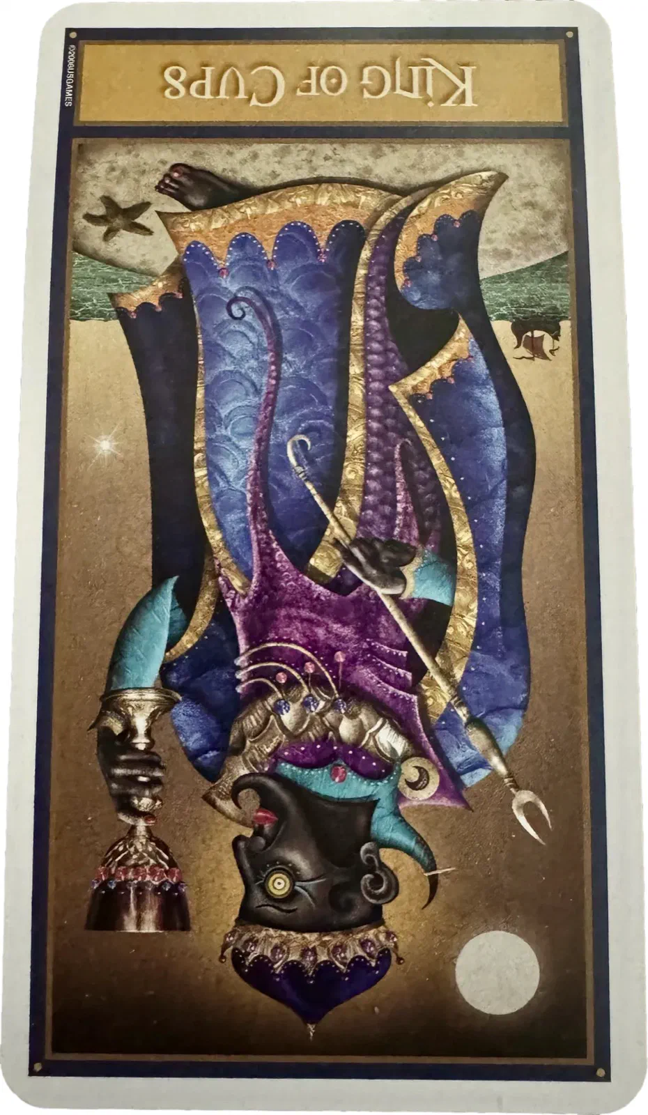 The King of Cups reversed. The card is upside down, so the shoreline that should sit behind the throne now runs across the top of the card above the king’s head — the sea is over him. His scaled blue, purple and gold robes fill almost the whole image; his crowned face sits at the very lowest point, turned away.
