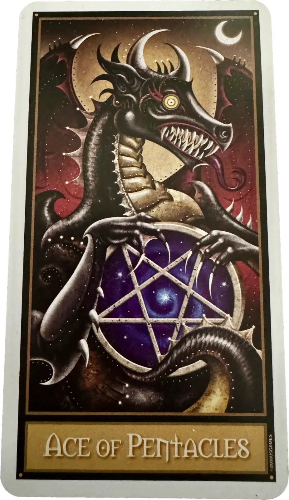 The Ace of Pentacles. A black horned dragon with its wings up and its teeth out is coiled around the pentacle, claws around the rim, holding the disc out toward the viewer and staring straight ahead.