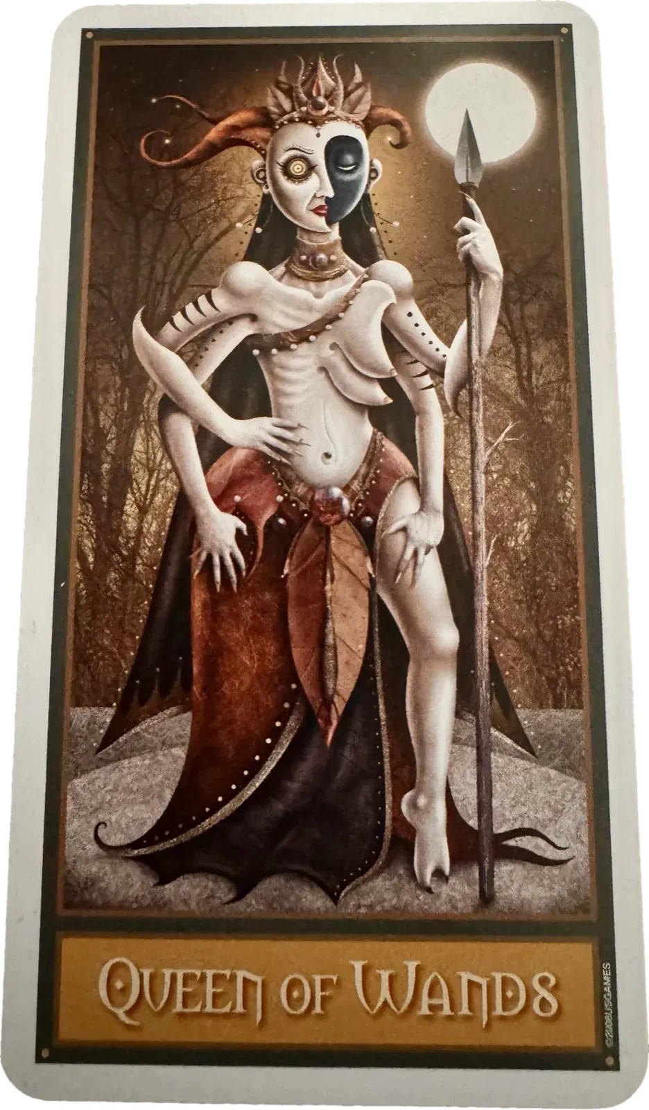 The Queen of Wands. A figure pale as chalk, bare from the waist up, crowned in something that reads as flames or antlers, holding a spear rather than a wand and standing under a full moon. One eye is dark and one is light.